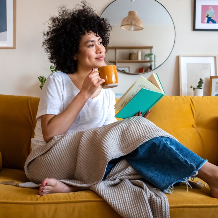 Pensive relaxed African american woman reading a book at home, drinking coffee sitting on the couch. Copy space. Pensive relaxed African american woman reading a book at home, drinking coffee sitting on the couch. Copy space. Lifestyle concept.