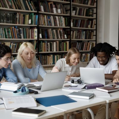 Group of attractive multi ethnic students studying together using laptop, prepare for exams seated at table in modern library. Cooperation, teamwork, higher education, modern tech usage, gen Z concept